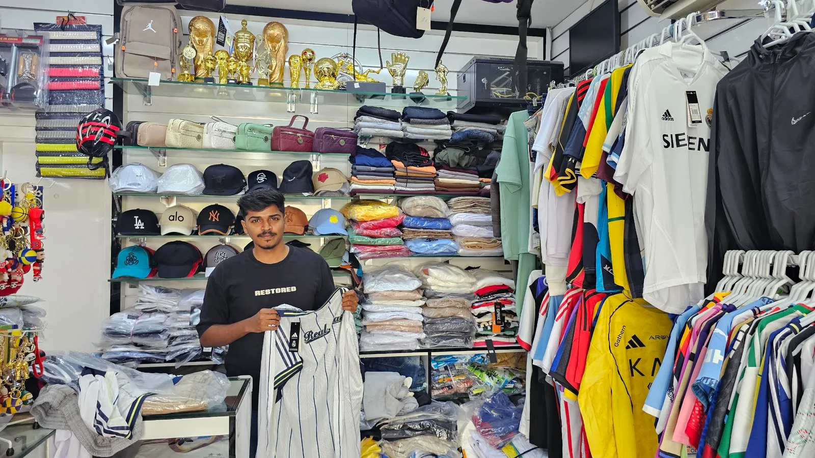 Inside Al Shamaa Al Baidaa: jerseys on rails, caps and bags on shelves, trophies above the counter